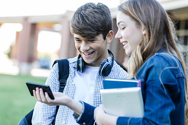 Two young students, boy and girl, smile while looking at a smartphone outdoors. They are holding books.