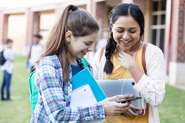 Friends, diverse ethnicities, sharing a laptop and phone, smiling with joy, holding books, outdoors.