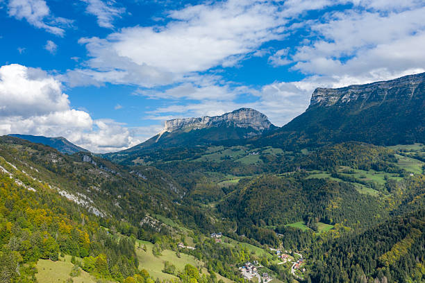 Wide aerial perspective of the Chartreuse Massif reveals dramatic limestone cliffs, rolling green valleys, and scattered forested slopes under a vibrant blue sky with scattered clouds. The scene captures the natural contours and lush vegetation of this iconic alpine region.