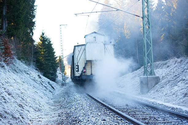 Rear view of a vintage steam train departing along a snowy railway track in winter forest. Thick steam cloud, frost-covered ground and railway infrastructure create a cold atmospheric scene. Concept of historic transport, travel, industrial heritage and motion