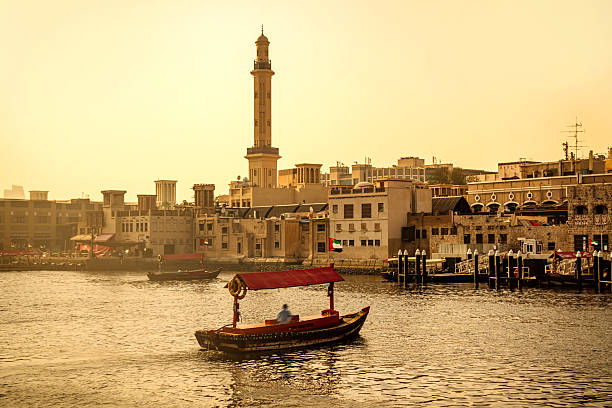 A warm, golden sunset over Dubai's harbor scene. A traditional wooden boat glides on calm water past historic buildings and a tall minaret, capturing maritime life and urban contrast.