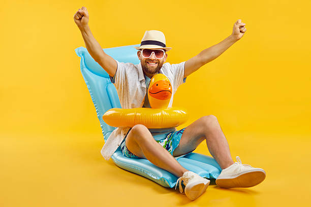 Happy funny excited man in sunglasses and hat sitting on inflatable mattress in duck rubber ring isolated on a studio yellow background. Overjoyed guy going to swim on a beach. Vacation concept.
