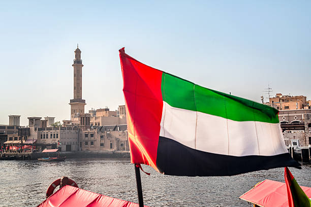 A vibrant UAE flag waves over Dubai's waterfront with traditional architecture and a tall minaret in the background. The scene conveys pride, travel, and the contrast between old and new.