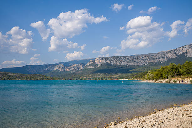 Vivid turquoise water stretches toward a rugged mountain range at Lac de Sainte-Croix, bordered by a stony shoreline and lush green forest under a sky dotted with puffy clouds.