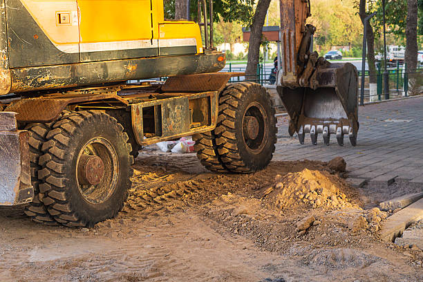 Construction site with excavator with its bucket lowered onto paving slab, rear and side view. Specialized equipment for excavation work, laying and replacing pipes, underground utilities. Work place.