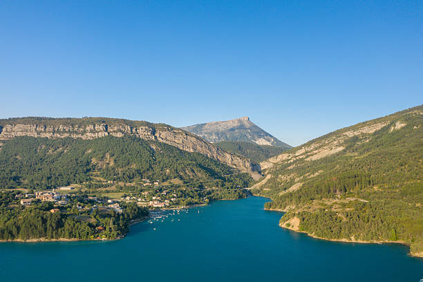 Aerial view of a lakeside village nestled among green hills and dramatic cliffs along the turquoise waters of Lac de Castillon, with a prominent mountain peak in the distance and clear summer skies overhead.