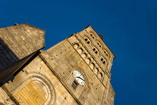 Warm evening light illuminates the stone clock tower and Romanesque arches of the abbey in La Charite sur Loire. The textured masonry stands out against a deep blue sky, highlighting historic architectural details.