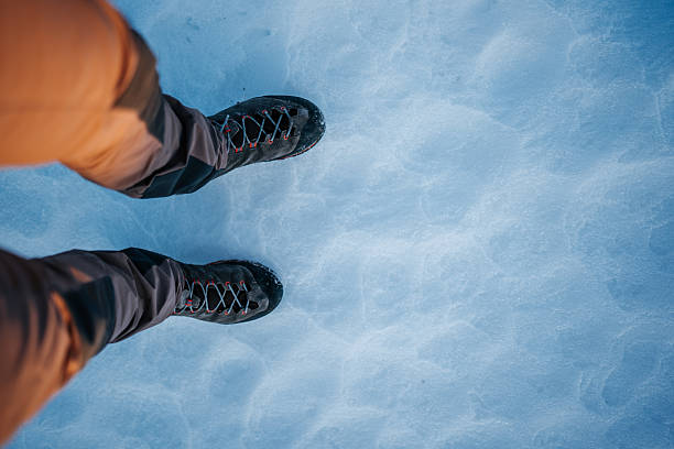 Top-down close-up of a hiker’s feet in durable dark winter boots with contrasting red and gray laces, standing on a compacted snow surface. The boots show signs of outdoor use, including light snow residue on the rubber toe caps and sidewalls. Natural low-angle daylight creates long, soft shadow lines across the textured snow, adding depth and a minimal, atmospheric aesthetic. The composition highlights winter travel, outdoor gear, seasonal conditions, and active lifestyle elements, suitable for stock themes including hiking, winter sports, cold-weather fashion, adventure tourism, and nature-based recreation.