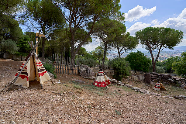 Small patterned teepees stand on a dry, wooded hillside among pine trees, overlooking a sprawling distant city beneath a clear blue sky with white clo