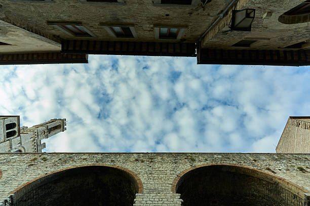 A unique low-angle view looking upward at the medieval stone architecture and arches framing dynamic white clouds sweeping across a bright blue sky, t
