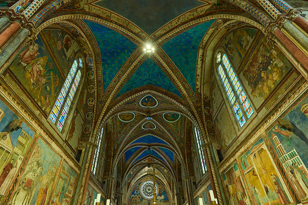 A stunning low-angle view of the highly decorated vaulted Gothic ceiling, frescoes, and colorful stained glass windows of the Basilica of Saint Francis in Assisi, Umbria, Italy.