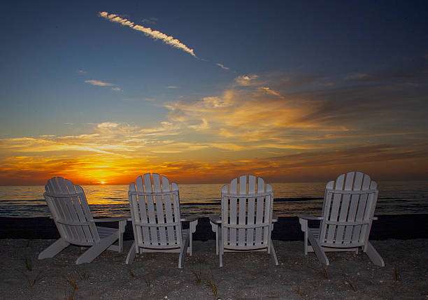 Four white Adirondack chairs face a vibrant sunset over the ocean, creating a serene and peaceful coastal scene on Captiva Island, Florida. The colors of the setting sun reflect on the water, while the sky features a spectrum of warm hues.