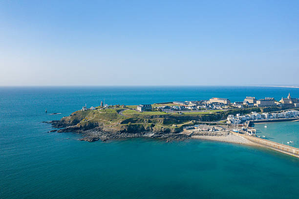 Aerial view of Pointe du Roc's grassy headland jutting into vivid turquoise water, with rugged rocks, a lighthouse, and coastal buildings under a clear blue sky. The scene is open and sunlit, evoking a sense of tranquility and space.