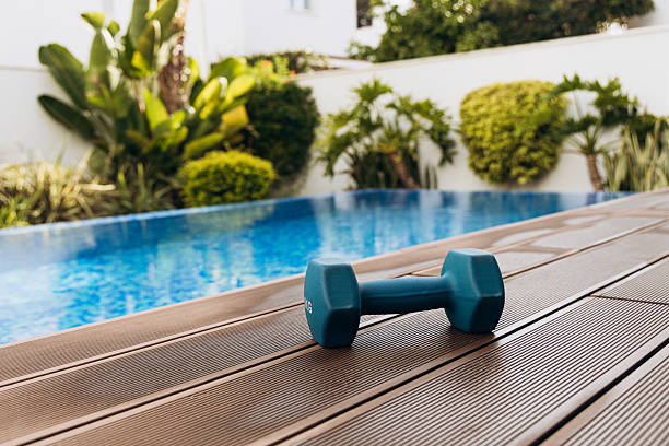 istock Blue dumbbell resting on wooden deck beside a swimming pool surrounded by lush greenery, showcasing fitness lifestyle and outdoor exercise environment 2254968995