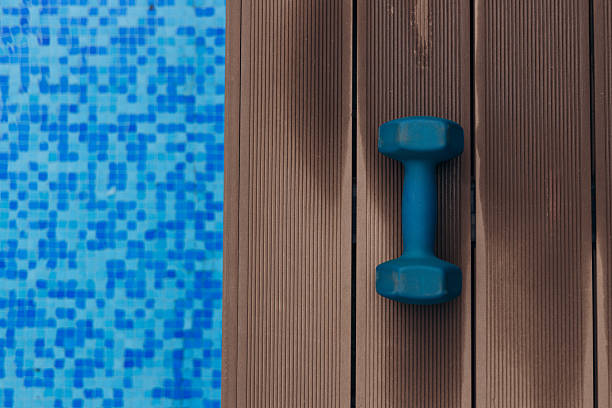 A blue dumbbell is placed on a wooden deck next to a sparkling swimming pool, showcasing a refreshing environment ideal for workouts and leisure activities