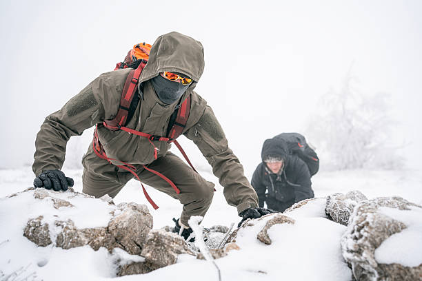 Two hikers in winter gear tackle a snow-covered rocky path, showcasing endurance, teamwork, and outdoor adventure in a cold, windy landscape.