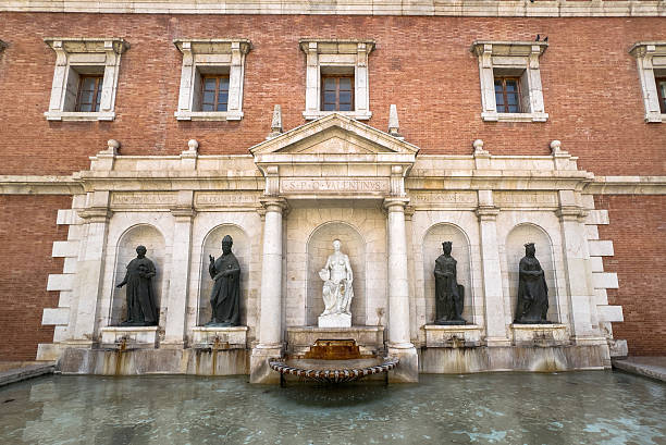 Historic Fuente de la plaza del Patriarca in Valencia, Spain, featuring four bronze statues flanking a central marble figure symbolizing the city, set against a grand red-brick facade.