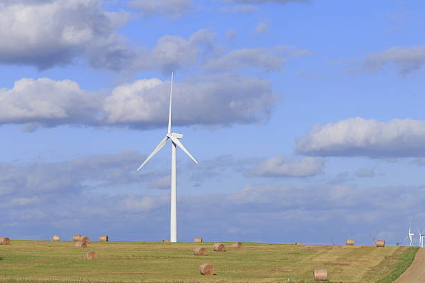 Electric windmill in a farm field with haybales, grass, and blue sky with white clouds east of Wilson Kansas USA out in the country.