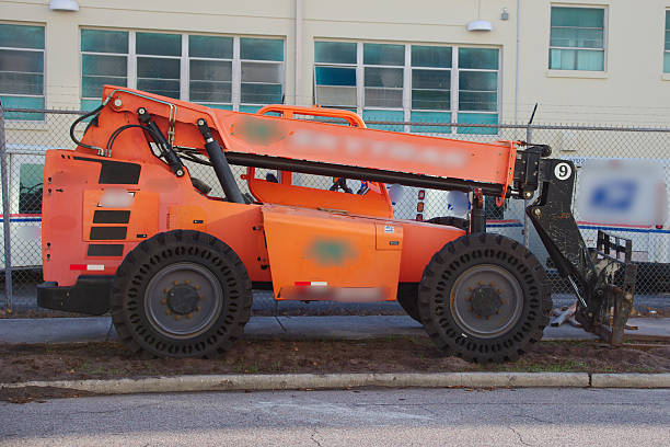 Vivid orange telehandler on a construction site beside a chain-link fence, with a modern building in the background. Suitable for industrial, construction, and logistics themes highlighting heavy machinery.