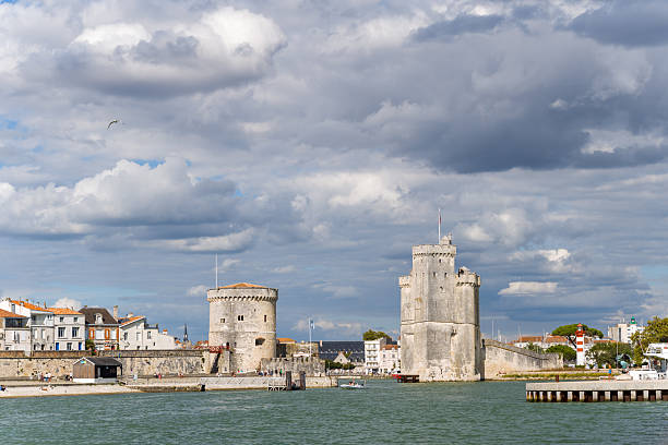 Iconic stone towers guard the entrance to the historic harbor of La Rochelle, Charente-Maritime, under a dramatic sky with layered clouds. The waterfront scene features fortified medieval architecture, old town buildings, and the calm Atlantic waters.