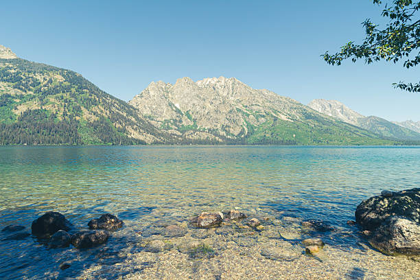 View across clear blue rippling waters of Jenny Lake from the rocky water's edge out towards th4e massive majestic snow-capped Grand Teton Mountain Range on a clear sunny day. Taken on a clear summer day in the Grand Teton National Park near Jenny Lake, a National Park located near Jackson Hole, Wyoming.