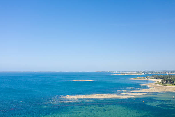 Expansive aerial view of a calm bay with visible sandbars and clear turquoise water near Saint Philibert, bordered by forest and distant coastline under a cloudless blue sky.