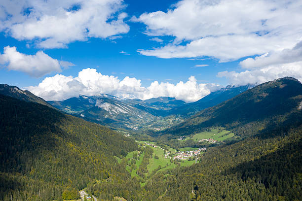 Expansive aerial perspective of a green alpine valley in the Chartreuse Massif, with dense forests, scattered villages, and dramatic peaks under a vivid blue sky and billowing clouds. The scene highlights the natural contours and lush vegetation of this iconic mountain region.