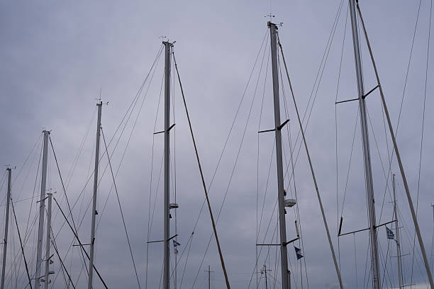 Sailboat masts standing in a marina with ropes and rigging against a cloudy sky. The image shows repeating vertical lines, nautical equipment and minimal maritime composition. This photo represents sailing, marine transport, harbor infrastructure and calm coastal atmosphere, suitable for travel, boating, yachting or abstract background concepts.