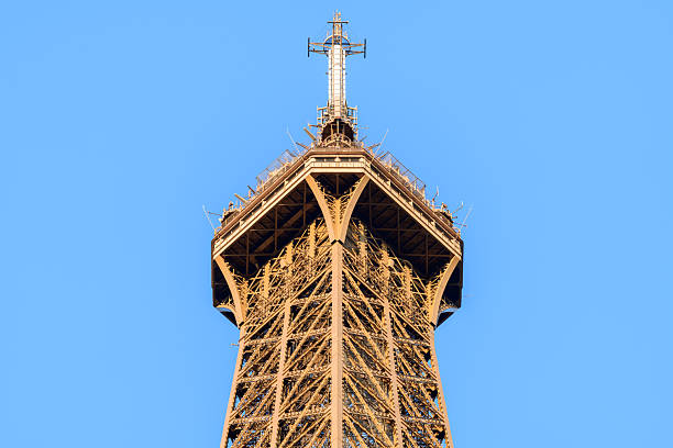Upward view of the Eiffel Tower's top section and antenna, showcasing detailed iron framework and geometric lines under bright daylight with a vivid blue sky backdrop.