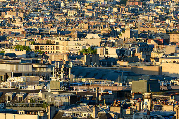Dense layers of Paris rooftops with varied architectural styles and rooftop structures, illuminated by warm evening light. The urban landscape features a rhythmic pattern of chimneys, antennas, and textured building facades.
