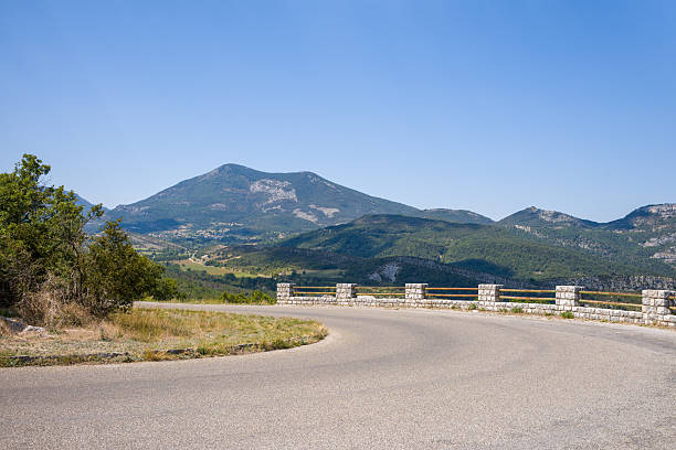 Empty asphalt road bends sharply beside a stone barrier, offering a panoramic view of forested hills and distant mountains under a clear blue sky in Alpes-de-Haute-Provence.