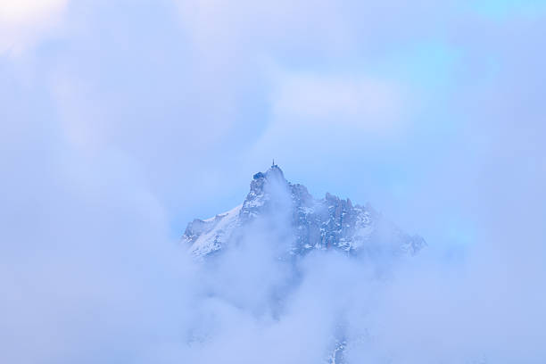 The snow-covered peak of Aiguille du Midi emerges through a veil of soft, blue-tinted clouds. Gentle light and thick fog create a tranquil, ethereal alpine scene.