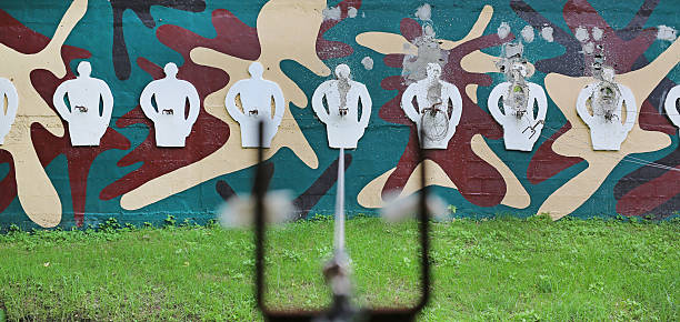 Perspective view from a shooting range through the sights of a weapon at white human-shaped targets on a camouflage wall.