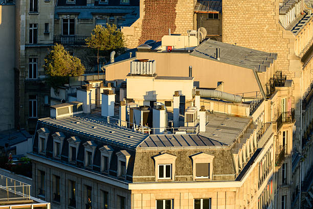 Close-up view of classic Paris rooftops featuring zinc surfaces, multiple chimneys, and dormer windows illuminated by warm evening sunlight. Architectural details and urban textures create a timeless city atmosphere.