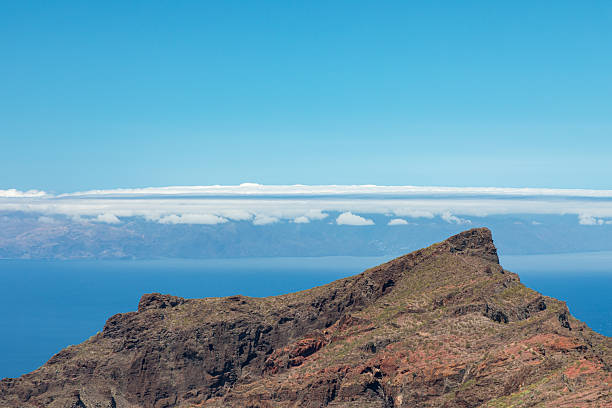 Rugged coastal cliff rises toward a bright blue sky, framing a calm sea and distant mountains. A striking natural landscape perfect for travel, nature, and adventure stock visuals.