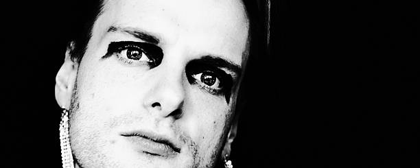 Close-up black and white portrait of a young man wearing expressive eye makeup and an earring. The direct gaze and dramatic lighting create a contemporary visual language exploring identity, self-expression, and emotional openness.