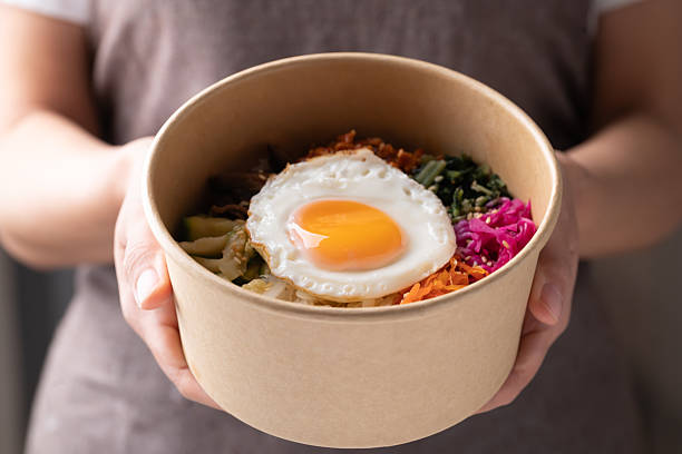 Bibimbap, Korean food, Cooked rice with vegetables, pork and egg in biodegrable bowl holding by woman hand