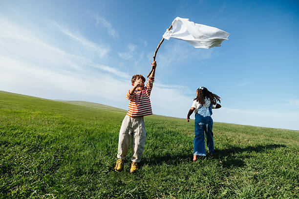 Two kids play in a grassy field while one waves a white flag, creating a lighthearted scene that evokes peace, unity, and hopeful childhood innocence.