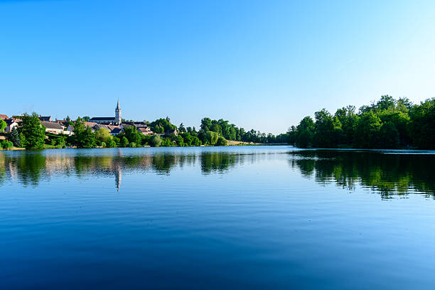 Still water reflects the charming village skyline and church spire of Dampierre en Burly, surrounded by lush green trees under a vibrant blue sky. The expansive lake and bright light evoke a sense of openness and tranquility.