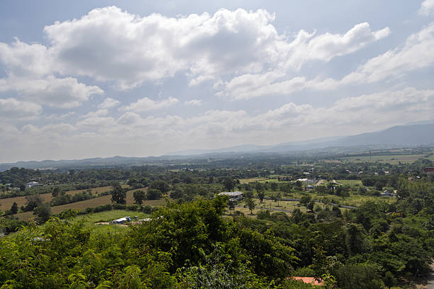 expansive landscape featuring lush green foliage in foreground, agricultural fields and houses in mid-distance, rolling hills and mountain range in background; bright blue sky dotted with large white cumulus clouds