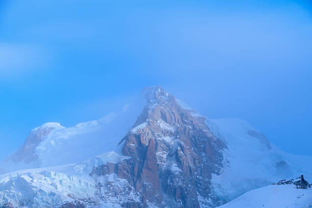 The jagged summit of Mont Maudit emerges through soft blue mist, with snow and ice clinging to rugged rock faces under tranquil early morning light.