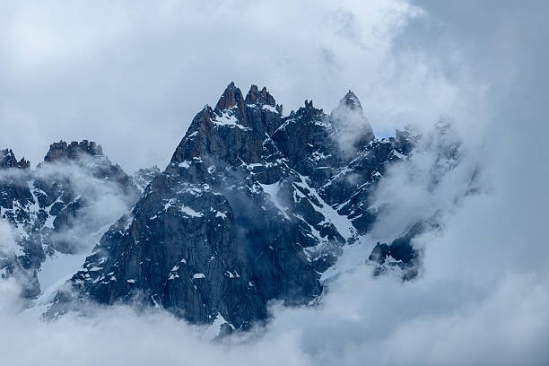 Jagged mountain summits rise through swirling clouds, with rugged rock faces and patches of snow creating a dramatic alpine scene in the French Alps.