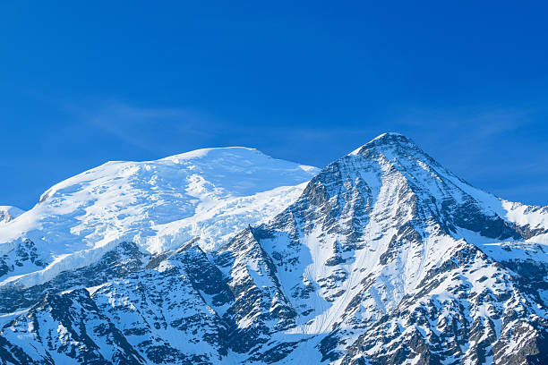 Snow-capped Mont Blanc and the rugged Aiguille du Gouter rise sharply against a vibrant blue sky, their icy slopes illuminated by bright sunlight in the French Alps.