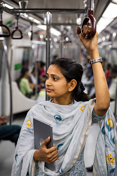 A woman stands inside a metro carriage holding an overhead strap with one hand while clutching a tablet to her chest with the other. She wears a light patterned dupatta and kurta, small gold earrings and a nose stud, and gazes thoughtfully toward the carriage interior. Passengers and seats in the background are softly blurred, highlighting the candid moment during an urban commute.