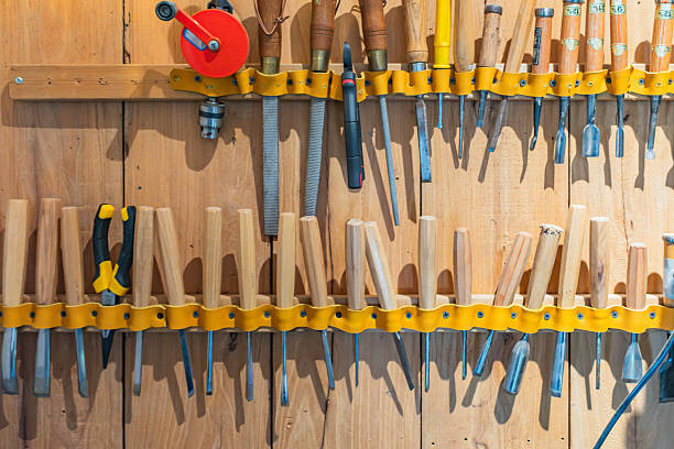 This image displays a neat assortment of carpentry and wood carving tools hanging on a light-colored wooden wall. The tools are secured in two rows by yellow undulating straps. The top row features a red manual hand drill, several metal files, and various chisels with wooden handles. The bottom row includes a pair of yellow-handled pliers and a long series of carving gouges and chisels of different shapes and sizes. Sharp shadows cast against the wood backing highlight the orderly arrangement and the texture of the tools.