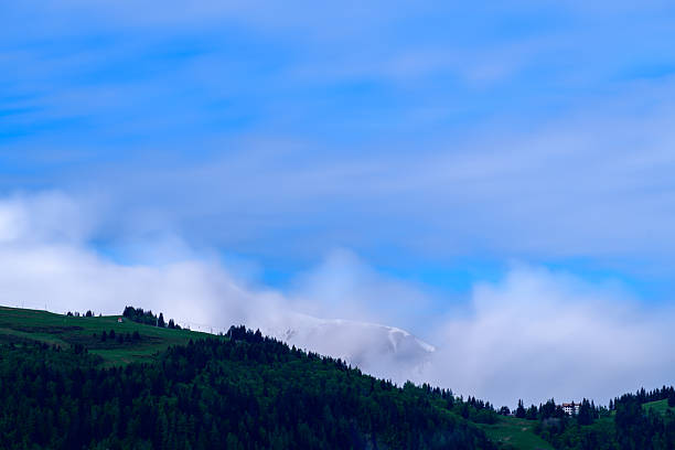 A distant snow-covered mountain summit is partially concealed by swirling clouds above lush green forested hills, with a vibrant blue sky overhead.