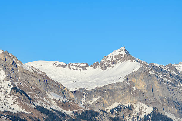 Sharp limestone summit rises above snowy alpine slopes in the Desert de Plate, highlighted by clear winter sunlight and a vivid blue sky.