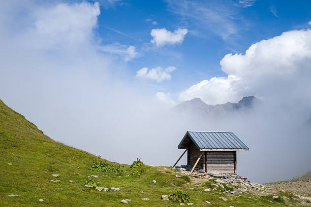 A solitary wooden shelter with a metal roof stands on a grassy hillside at Col du Bonhomme, surrounded by drifting fog and distant peaks under a vivid blue sky.