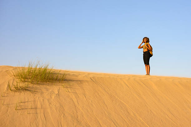 A lone hiker stands on a wide, wind-sculpted sand dune under a vivid blue sky. Wearing a red tank top, olive shorts and sturdy hiking boots, the person looks through binoculars toward the horizon. Sparse patches of grass dot the pale sand slopes, and a few fluffy white clouds add contrast to the deep sky. The scene conveys bright midday light, open space and a sense of exploration.