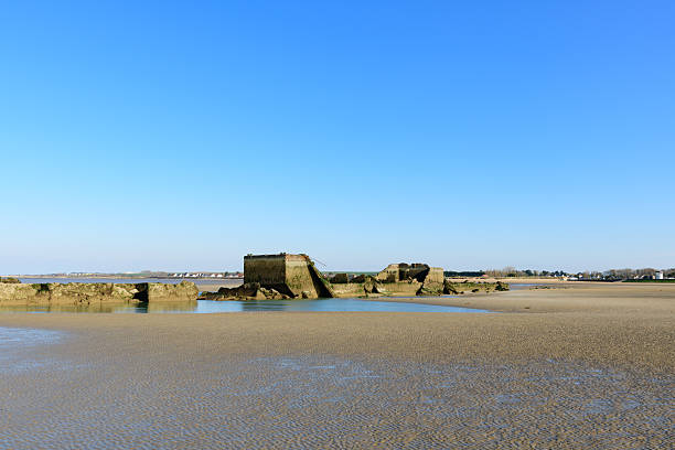 Weathered concrete remnants of an old harbor rest on vast sandy flats at low tide near Asnelles, Normandy, beneath a clear blue sky with distant village houses on the horizon.